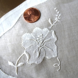 May include: White embroidered floral design on a white linen handkerchief. The design features a large flower with a stem and leaves.