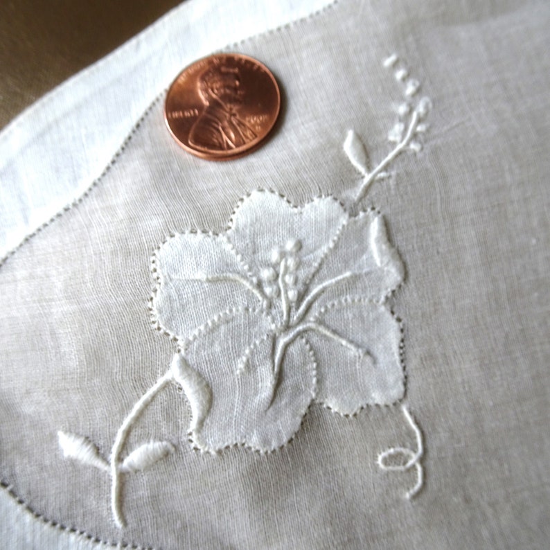 May include: White embroidered floral design on a white linen handkerchief. The design features a large flower with a stem and leaves.