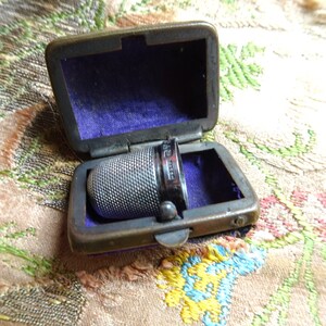 May include: A silver thimble with a textured surface is inside a small, rectangular, brass case lined with purple velvet. The case has a hinged lid.