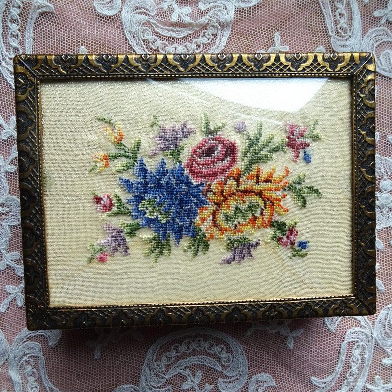 May include: A vintage floral embroidery framed in a gold-toned metal frame. The embroidery features a bouquet of colorful flowers, including pink, yellow, blue, and green.