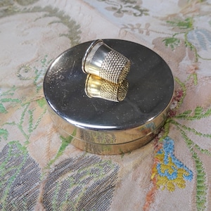 May include: A gold metal thimble sits on top of a round, gold metal box. The box is closed and has a smooth, shiny surface.
