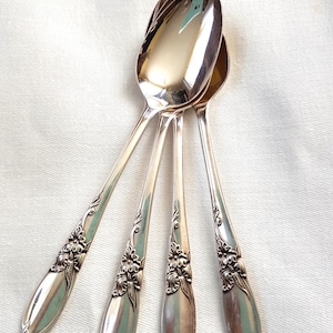 May include: Four silver spoons with ornate floral detailing on the handles, arranged on a white cloth. The spoons have a polished, reflective surface. The spoons are angled, displaying their elegant design and shape.