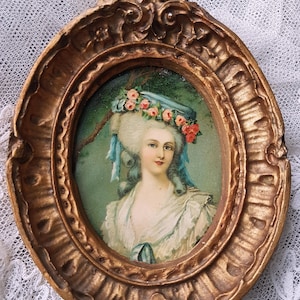 Puede incluir: Un marco ovalado de color dorado antiguo rodea un retrato de una mujer con un vestido blanco y un sombrero azul adornado con flores. El retrato tiene un fondo verde y el marco tiene detalles ornamentados.