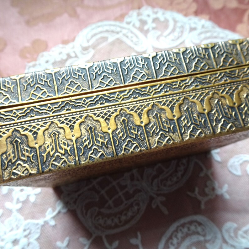 May include: A gold-colored box with intricate black and gold patterned designs. The box has a hinged lid and is decorated with a repeating pattern of arches and geometric shapes.