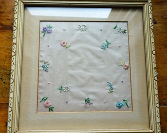 Op de afbeelding: Een ingelijst geborduurd zakdoek met een witte achtergrond en kleurrijke bloemenborduurwerk. De lijst is lichtbeige met een decoratieve rand.