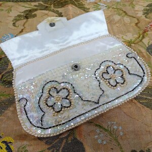 May include: A white satin clutch purse with a beaded and sequined design. The purse has a floral motif and a pearl trim.