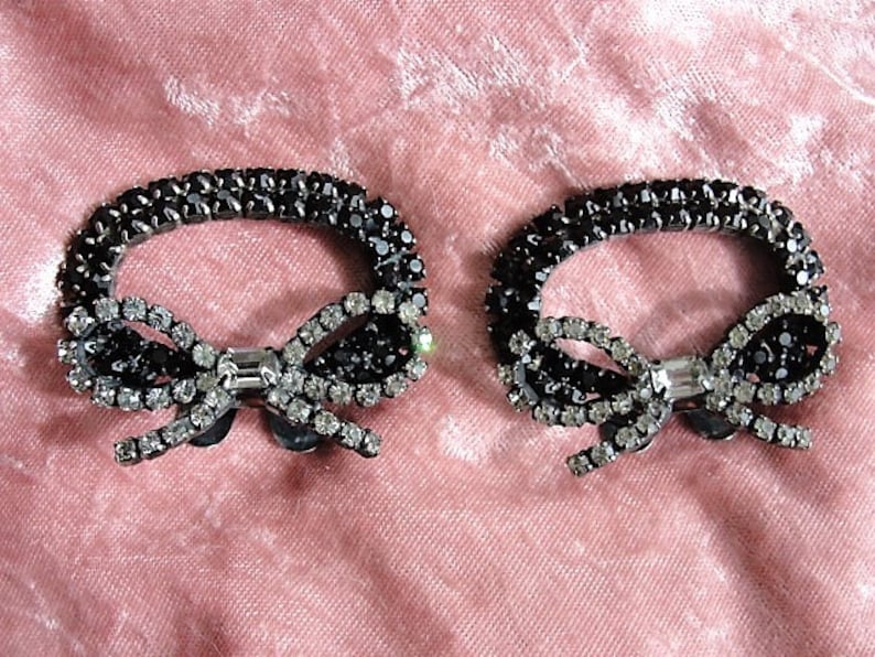 K&ouml;nnte beinhalten: Ein Paar schwarze und silberne Strass-Schuhclips in Form einer Schleife. Die Clips sind aus Metall und haben eine gl&auml;nzende Oberfl&auml;che.