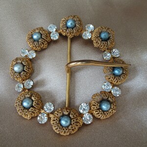 May include: A gold-toned belt buckle with a decorative design of blue faux pearls and clear rhinestones set in a circular pattern.