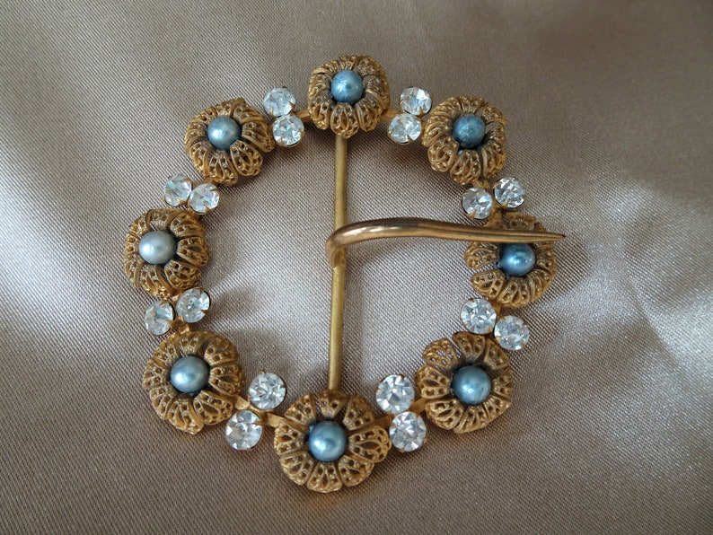 May include: A gold-toned belt buckle with a decorative design of blue faux pearls and clear rhinestones set in a circular pattern.