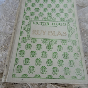 Pode incluir: Um livro vintage intitulado "Ruy Blas" de Victor Hugo. A capa do livro &eacute; branco sujo com um padr&atilde;o verde repetido e texto verde. O livro est&aacute; ligeiramente aberto, revelando as p&aacute;ginas envelhecidas.