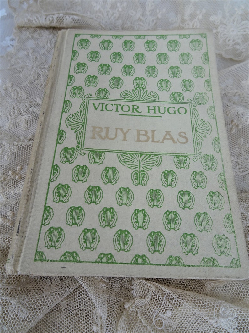 Pode incluir: Um livro vintage intitulado "Ruy Blas" de Victor Hugo. A capa do livro &eacute; branco sujo com um padr&atilde;o verde repetido e texto verde. O livro est&aacute; ligeiramente aberto, revelando as p&aacute;ginas envelhecidas.