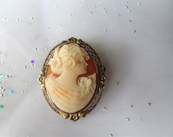 May include: A cameo brooch featuring a carved portrait of a woman with a flower in her hair, set in a gold-toned filigree frame.