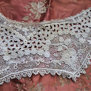 Puede incluir: Un delicado cuello de encaje blanco tejido a crochet con un borde festoneado. El encaje presenta intrincados patrones florales y peque&ntilde;os detalles de cuentas.