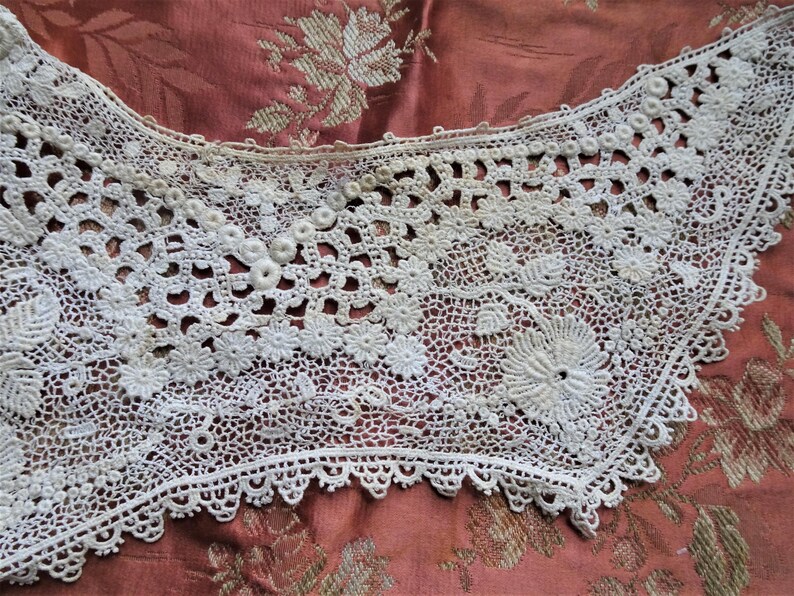 Puede incluir: Un delicado cuello de encaje blanco tejido a crochet con un borde festoneado. El encaje presenta intrincados patrones florales y peque&ntilde;os detalles de cuentas.