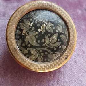 Pode incluir: Um botão redondo dourado com um design floral preto e prateado. O botão tem uma borda texturizada.