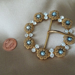 May include: A gold-toned belt buckle with a circular design. The buckle is decorated with blue and white rhinestones and faux pearls.