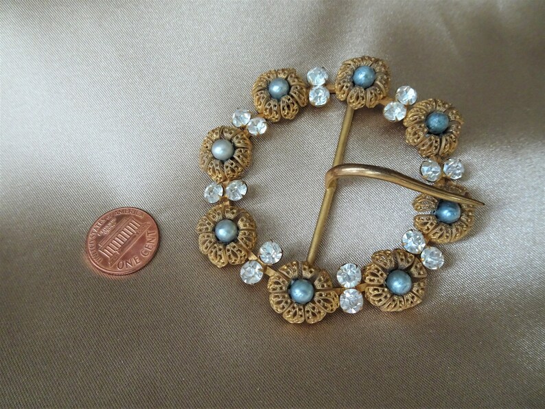 May include: A gold-toned belt buckle with a circular design. The buckle is decorated with blue and white rhinestones and faux pearls.
