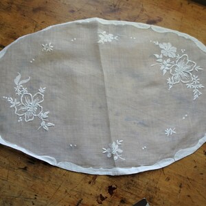 May include: White oval-shaped doily with delicate white floral embroidery. The doily has a scalloped edge and is made of a sheer fabric.