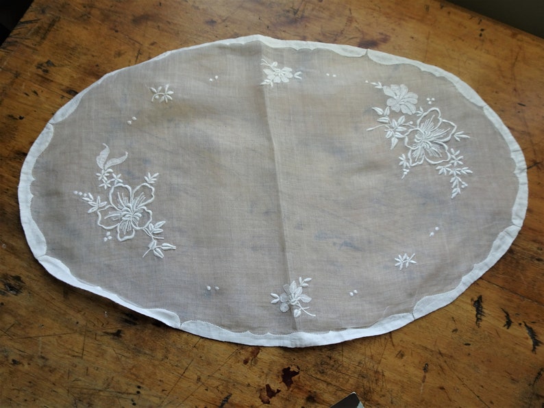 May include: White oval-shaped doily with delicate white floral embroidery. The doily has a scalloped edge and is made of a sheer fabric.