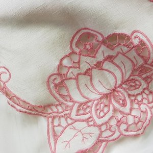 May include: A white linen tablecloth with a pink embroidered floral design. The embroidery features a large flower with intricate details and smaller leaves.