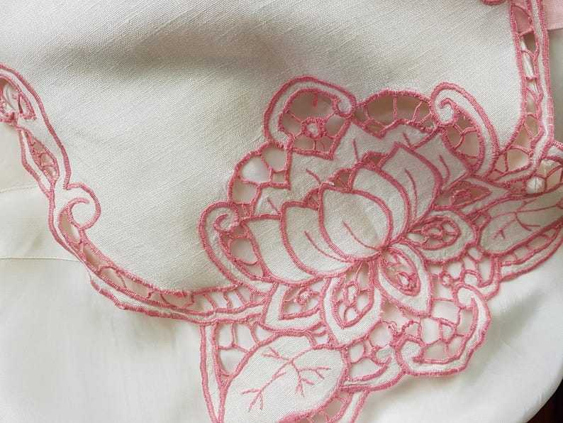 May include: A white linen tablecloth with a pink embroidered floral design. The embroidery features a large flower with intricate details and smaller leaves.