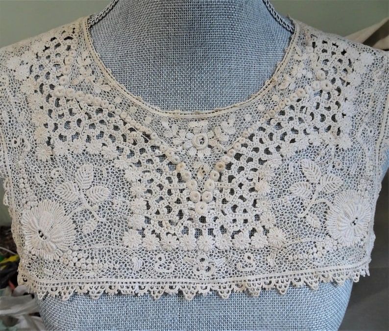 Puede incluir: Un delicado cuello de encaje blanco con borde festoneado. El cuello est&aacute; hecho de intrincado trabajo de ganchillo y presenta un dise&ntilde;o floral.