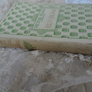 Pode incluir: Um livro antigo, "Ruy Blas" de Victor Hugo, com capa creme e elementos decorativos verdes. O livro est&aacute; sobre renda branca. O t&iacute;tulo e o autor s&atilde;o vis&iacute;veis na capa.