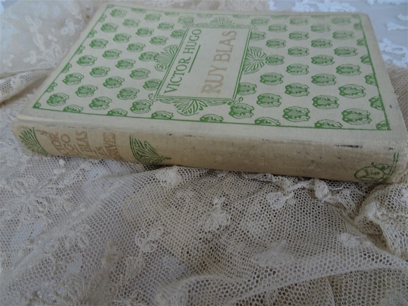 Pode incluir: Um livro antigo, "Ruy Blas" de Victor Hugo, com capa creme e elementos decorativos verdes. O livro est&aacute; sobre renda branca. O t&iacute;tulo e o autor s&atilde;o vis&iacute;veis na capa.
