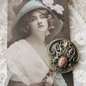 May include: A vintage postcard featuring a woman wearing a light blue hat with pink flowers. She is wearing a white dress with a floral pattern. A silver and purple cameo brooch is pinned to her dress.