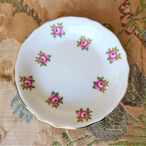 Puede incluir: Un plato de porcelana blanca con borde dorado y un delicado patr&oacute;n floral de rosas rosas con hojas verdes.