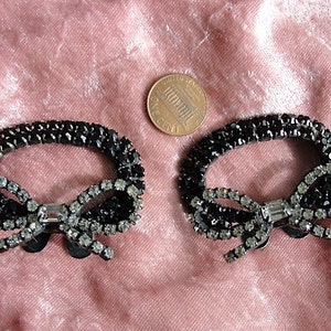 K&ouml;nnte beinhalten: Zwei schwarze und silberne Strass-Haarspangen in Schleifenform. Jede Spange hat eine schwarze Schleife mit einem silbernen Strassstein in der Mitte und einem schwarzen ovalen Rahmen.