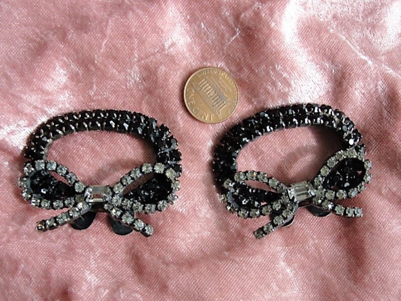 K&ouml;nnte beinhalten: Zwei schwarze und silberne Strass-Haarspangen in Schleifenform. Jede Spange hat eine schwarze Schleife mit einem silbernen Strassstein in der Mitte und einem schwarzen ovalen Rahmen.