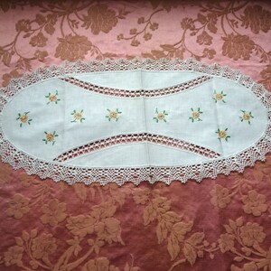 Puede incluir: Un camino de mesa ovalado blanco con un borde de encaje de ganchillo y flores amarillas bordadas con hojas verdes.