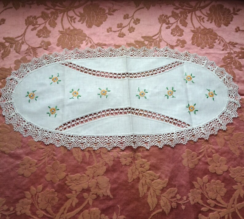 Puede incluir: Un camino de mesa ovalado blanco con un borde de encaje de ganchillo y flores amarillas bordadas con hojas verdes.