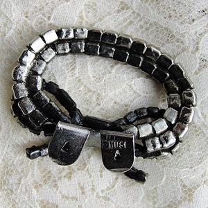 K&ouml;nnte beinhalten: Ein silbernes und schwarzes Armband mit einer doppelten Reihe quadratischer Perlen. Das Armband hat einen Verschluss mit der Gravur "OMUST".