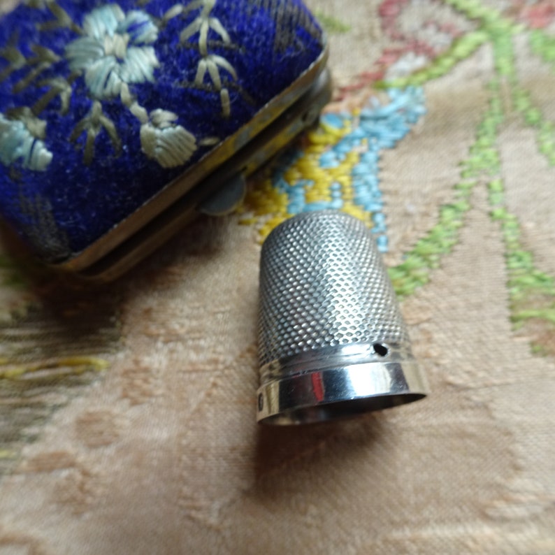 May include: A silver thimble with a textured surface and a small hole at the top. The thimble is sitting on a fabric surface with a floral pattern.