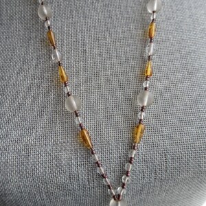Peut inclure: Un long collier avec des perles de verre transparentes et ambr&eacute;es altern&eacute;es. Le collier est sur un fond de tissu gris.