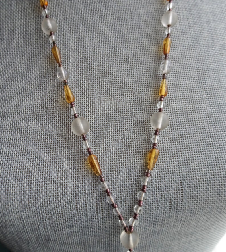 Peut inclure: Un long collier avec des perles de verre transparentes et ambr&eacute;es altern&eacute;es. Le collier est sur un fond de tissu gris.