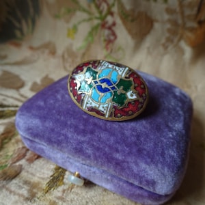 May include: An oval-shaped brooch with a colorful enamel design. The design features a central blue and green motif surrounded by red and white accents. The brooch is sitting on a purple velvet box.