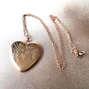Op de afbeelding: Een hartvormige medaillonhanger in roségoudkleur, hangend aan een bijpassende fijne schakelketting. De voorzijde van het medaillon is voorzien van een gedetailleerd gegraveerd bloem- en schildmotief met een parelrand langs de rand.