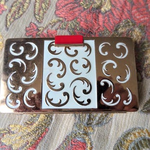 Op de afbeelding: Een gouden en witte compact met een decoratief ontwerp van draaiende patronen. De compact heeft een rode accent op de bovenkant.