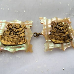 May include: A pair of antique mother-of-pearl and brass dress clips. Each clip features a cherub figure with wings, holding a spinning wheel. The clips are connected by a metal chain.