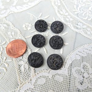 May include: Six small, round, black buttons featuring a raised, textured floral pattern. A copper-coloured one-cent coin is positioned to the left of the buttons for scale, providing a clear visual reference for their size.