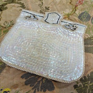 May include: A white iridescent sequined purse with a black beaded trim. The purse has a small, black, beaded flower design on the front.