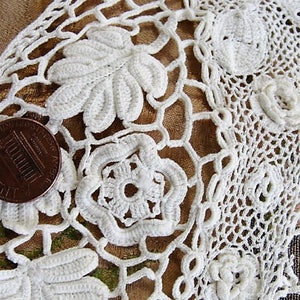 May include: White crocheted lace trim with a floral design. The lace is delicate and intricate, with a repeating pattern of flowers and leaves. A US penny is included for scale.