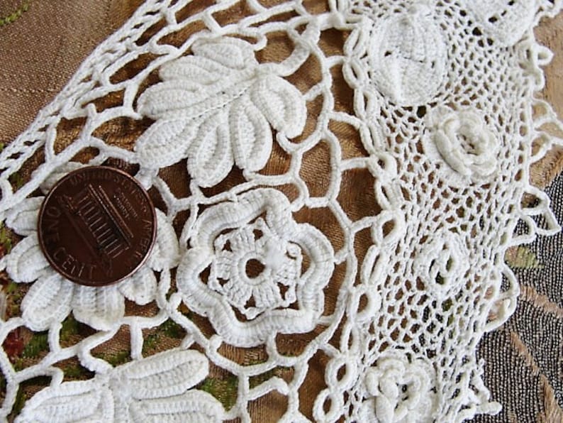 May include: White crocheted lace trim with a floral design. The lace is delicate and intricate, with a repeating pattern of flowers and leaves. A US penny is included for scale.