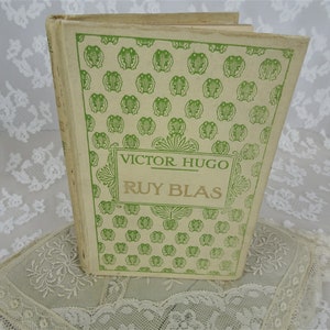 Pode incluir: Um livro vintage intitulado "Ruy Blas" de Victor Hugo. A capa do livro &eacute; creme com um padr&atilde;o de r&atilde;s verdes e texto. O livro est&aacute; sobre uma superf&iacute;cie de renda branca.