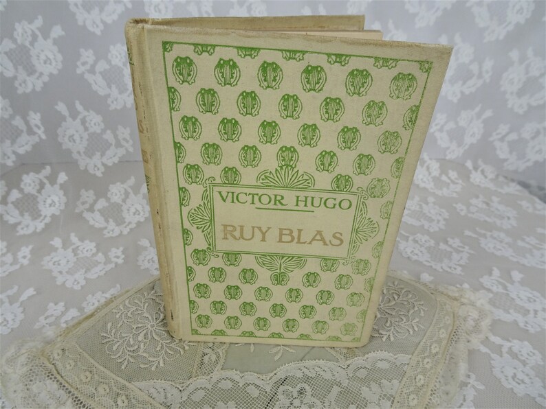 Pode incluir: Um livro vintage intitulado "Ruy Blas" de Victor Hugo. A capa do livro &eacute; creme com um padr&atilde;o de r&atilde;s verdes e texto. O livro est&aacute; sobre uma superf&iacute;cie de renda branca.