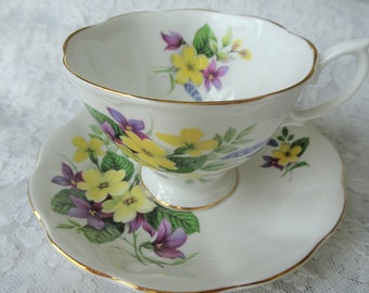 May include: A white teacup and saucer with a floral pattern. The pattern features yellow, purple, and green flowers. The cup and saucer have a gold trim.