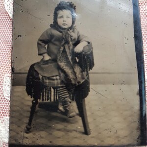 May include: A black and white tintype photograph of a young child wearing a dark coat and a bonnet, sitting on a chair with a fringed seat.